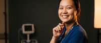 Young Asian female healthcare professional in blue scrubs smiling confidently in modern medical facility with equipment in background for healthcare marketing.
