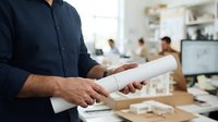 Male architect or contractor in dark navy shirt holding rolled white blueprint plans in modern office workspace with colleagues collaborating in background.