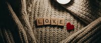 Wooden letter tiles spelling LOVE with a red felt heart on a cozy knit sweater, warm romantic mood for Valentine's Day cards and gifts.