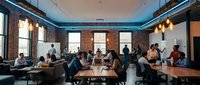 Busy modern coworking space with exposed brick walls and large windows. Diverse professionals working at wooden tables in open office environment.