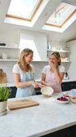 Two young Caucasian women in casual aprons prepare food together in a bright, modern kitchen with skylights, marble countertops, and open shelving. Perfect for cooking, lifestyle, and home content.