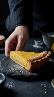 Caucasian male pastry chef hand placing a lemon tart slice on dark slate plate, dusted with powdered sugar, moody dark food styling.