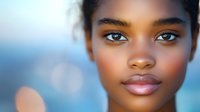 Young African American woman with natural makeup and glowing skin against blurred blue background with soft bokeh lighting effect and gentle expression.