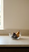 Fresh figs in clear glass bowl on marble countertop with neutral beige wall background, minimalist kitchen still life arrangement for home decor and culinary content.