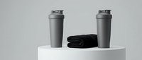 Two gray shaker bottles and black towel on white cylindrical podium against neutral background, suitable for fitness, gym, sports nutrition, and wellness product marketing.
