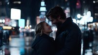 Romantic couple embracing in rain at night, city lights bokeh background creates atmospheric mood. Young blonde woman and dark-haired man share intimate moment outdoors.