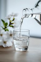 Pouring pure water from glass bottle into transparent drinking glass with white flowers in background, capturing refreshing splash and bubbles on wooden surface.