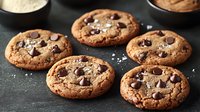 Fresh baked chocolate chip cookies with sea salt flakes on dark background, warm homemade dessert with melted chocolate pieces and golden brown edges.