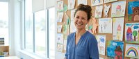 Happy female teacher smiling in bright classroom with colorful student artwork displayed on wall behind her during school day.