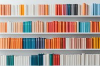 Books arranged by color on white shelves creating rainbow pattern. Minimalist library interior with hardcover volumes in coral, turquoise, and neutral tones.