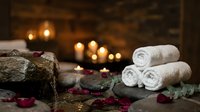 Spa wellness setup with rolled white towels, volcanic stones, rose petals and candles creating relaxing atmosphere for massage therapy treatments.