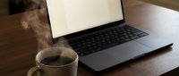 Steaming coffee mug beside an open laptop with blank screen on wooden desk, cozy remote work setup with warm morning atmosphere.