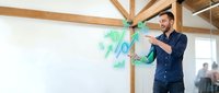 Man in casual business attire pointing at green percentage symbols floating in air, representing financial growth, discount rates, or profit margins in modern office setting.