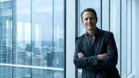 Confident middle-aged Caucasian businessman with crossed arms standing by office windows overlooking city skyline for corporate leadership content.