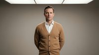 Professional middle-aged Caucasian businessman in beige cardigan and white shirt standing confidently in modern office environment for corporate marketing.