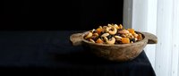Mixed nuts and dried fruits in rustic wooden bowl on dark surface near window with natural lighting for healthy snacking and cooking ingredients.