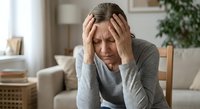 Stressed middle-aged Caucasian woman holding head in hands experiencing headache or mental fatigue in modern living room interior.