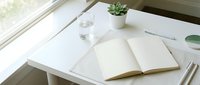 Clean minimalist workspace with open notebook, glass of water and small succulent plant on white desk by window for productivity and organization.