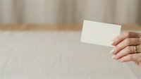 Close-up of a woman's hand holding a blank white card against a neutral beige linen background, suitable for business branding and marketing.