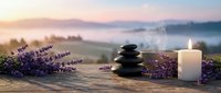 Zen spa composition with stacked black stones, burning candle and fresh lavender flowers on wooden surface against misty mountain landscape at sunrise.