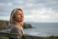 Mature Caucasian woman with gray hair enjoying peaceful moment by ocean coastline during golden hour sunset for wellness and retirement lifestyle concepts.