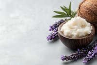 Organic coconut oil in coconut shell bowl with fresh lavender flowers and tropical leaves on light gray concrete background, spa and wellness concept.