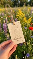 Hand holding seed paper tag labeled wildflower mix with twine in blooming meadow filled with purple lupine, yellow goldenrod, red poppies and blue cornflowers for gardening and eco-friendly gifting.