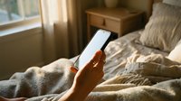 Morning routine with smartphone in cozy bedroom. Hand holding mobile device with blank screen for mockup. Soft natural light creates warm atmosphere for relaxation.