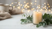 Cozy winter home decor with burning candle, eucalyptus leaves, warm knitted blanket and coffee cup. Soft bokeh lights create peaceful hygge atmosphere.