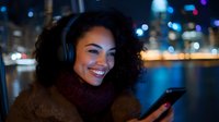 Young African American woman with curly hair wearing headphones smiles while using smartphone at night, city lights and waterfront in background, warm ambient lighting.