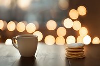 White coffee cup and stack of pancakes on wooden table against blurred warm bokeh lights background creating cozy morning atmosphere.