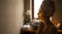Young African woman with towel wrapped hair relaxing in spa bathroom with candles and orchid flower for wellness skincare routine and self-care ritual.