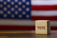 Wooden cube with vote text against blurred American flag background on wooden table, representing democracy, election and political concept.