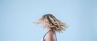 Young blonde woman with flowing hair in motion against clean blue background, expressing freedom and joy in casual summer style.