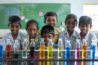 South Asian students smile behind colorful chemistry experiment test tubes in classroom laboratory setting, demonstrating hands-on science education and learning.