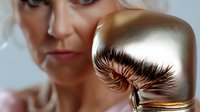 Dramatic close-up of golden boxing glove with long false eyelashes, blending fashion and sport concepts against blurred female athlete background.