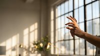 Hand reaching toward sunlight streaming through industrial window in modern loft space with warm natural lighting and soft shadows creating peaceful atmosphere.