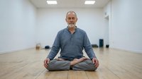 Mature man with gray beard, Caucasian ethnicity, sitting in lotus position on wooden floor in empty studio space. Wearing casual blue-gray long-sleeve shirt. Practicing meditation or yoga indoors.