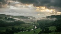 Misty valley landscape with winding river, rolling green hills, and dramatic cloudy sky at sunrise or sunset. Scenic nature background.