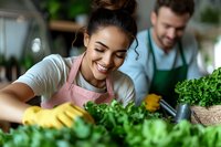 Young mixed race woman in pink apron and yellow gloves smiling while sorting fresh green vegetables at restaurant kitchen, male coworker in background.