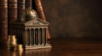 Bronze miniature bank building with classical columns surrounded by stacked coins on wooden surface with vintage books in background for financial concepts.
