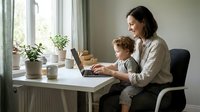 Mother and young son working together on laptop computer at home office desk by window with plants creating cozy family workspace atmosphere.
