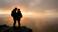 Two hikers silhouetted against golden sunrise on mountain peak, embracing after successful climb with backpacks and trekking gear overlooking misty valleys.