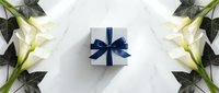 White gift box with blue ribbon surrounded by elegant white roses and green leaves on marble background for special occasions and celebrations.