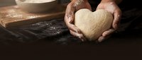 Hands holding heart-shaped dough in rustic kitchen setting with flour and baking ingredients on wooden surface for homemade bread making and cooking.