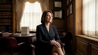 Mature woman, Caucasian ethnicity, wearing professional black blazer, sitting in traditional law office with wooden paneling, bookshelves, and natural window light. Suitable for legal, corporate, and professional business concepts.