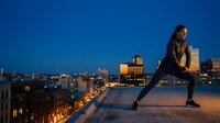 Young African American woman doing stretching exercise on urban rooftop at dusk with illuminated city skyline background for fitness and wellness concepts.