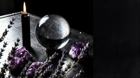 Crystal ball with burning black candle, amethyst crystals and dried lavender on dark surface, mystic fortune telling and spiritual ritual setup.