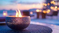 Glowing decorative metal bowl with flame against blurred evening cityscape background creates cozy atmosphere with warm bokeh lights.