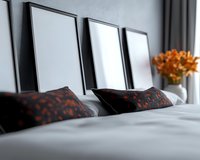 Modern bedroom interior with empty black frames above bed, white bedding, patterned orange and black decorative pillows, and orange flowers in vase.
